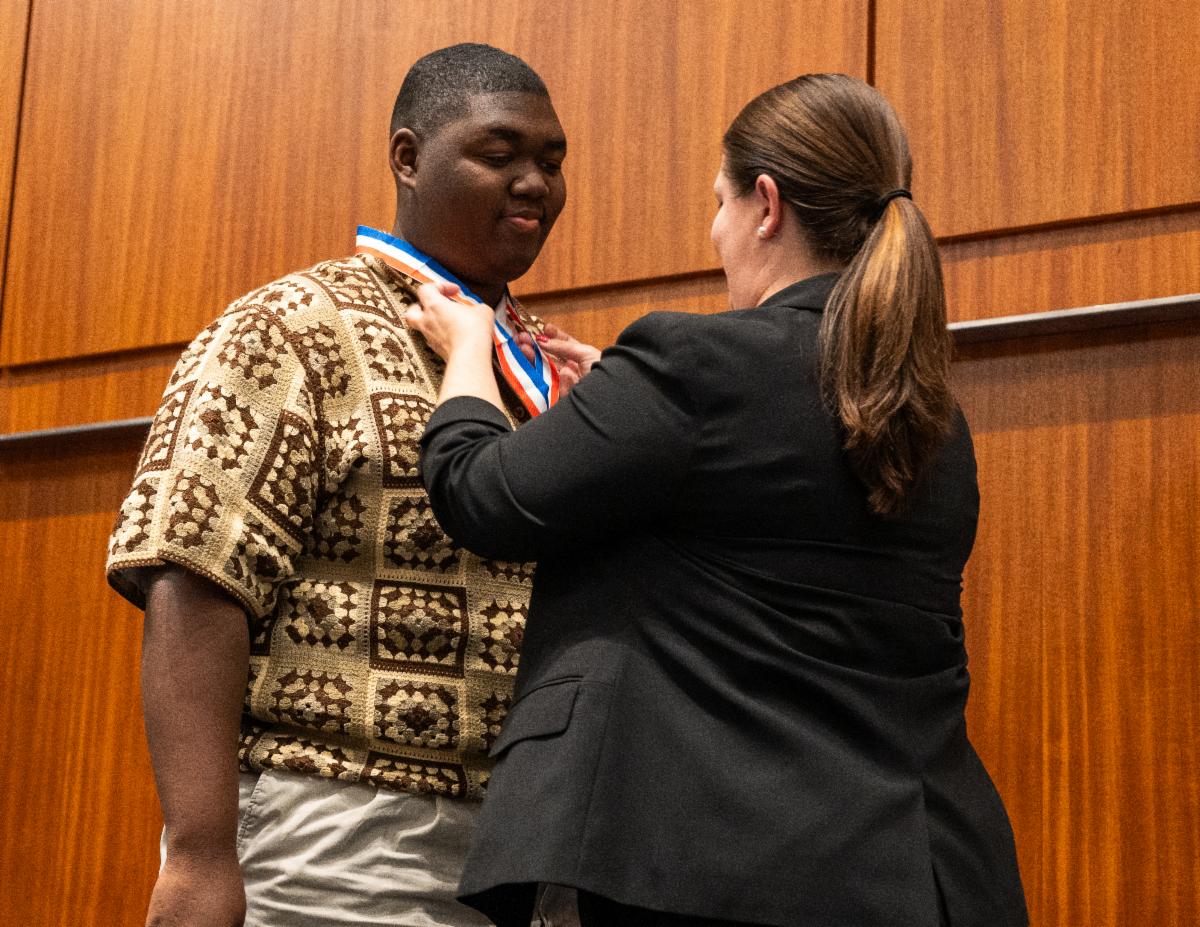 A woman places a medal around a young man's neck in a formal indoor setting with wooden walls.