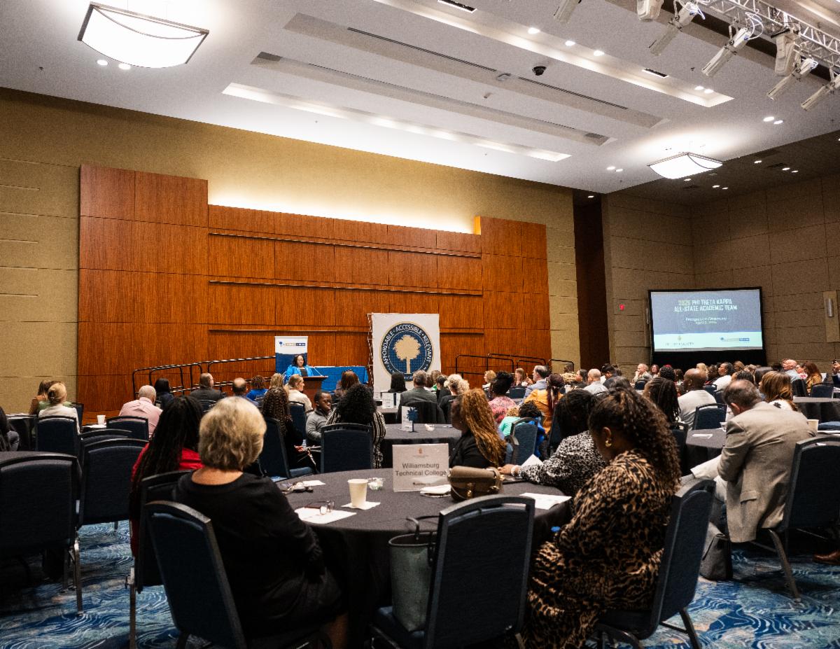 People attend a conference or seminar in a large, well-lit room, sitting around tables and facing a stage with speakers and a presentation screen.