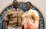 A young man wearing a medal stands next to a woman, both smiling, in front of a large circular emblem or logo.