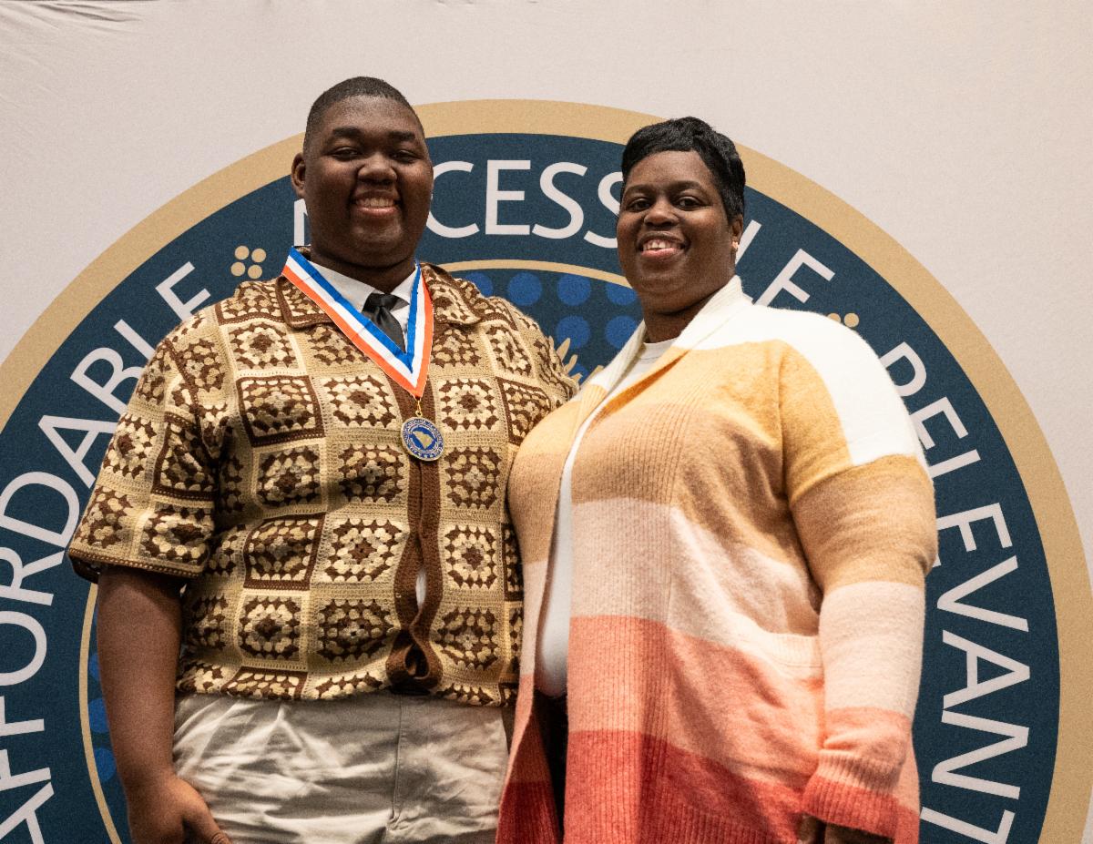 A young man wearing a medal stands next to a woman, both smiling, in front of a large circular emblem or logo.