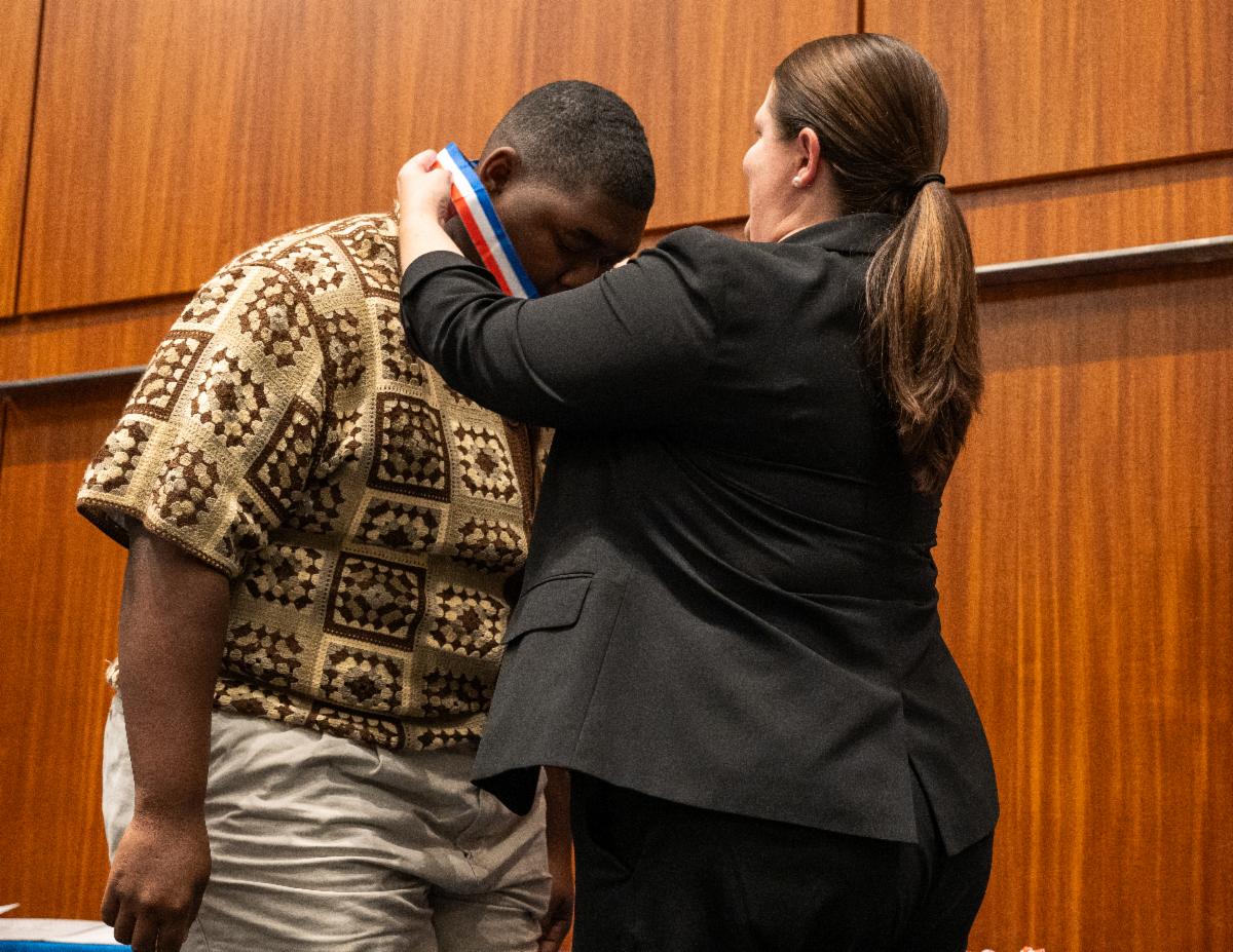 A woman in a black suit places a medal around a man's neck during an indoor ceremony with wooden panel walls.