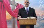 Man in suit stands at wooden podium with open Bible, large cross with red cloth and scenic sky in background.