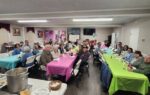 People sitting at long tables with colorful tablecloths in a community hall, sharing a meal and engaging in conversation.