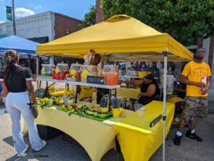 People at a street market stand with yellow decorations selling lemonade and fruit drinks under a yellow canopy on a sunny day.