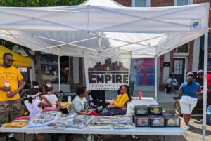 Outdoor market booth with "The Empire" banner, two women seated behind a table displaying shirts and boxes, people and children standing nearby, and a brick building in the background.