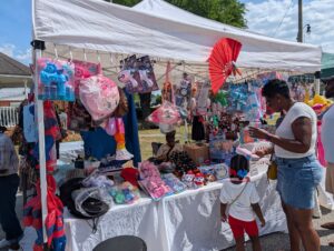 A woman and child browse toys and colorful items at an outdoor market stall under a white canopy on a sunny day.