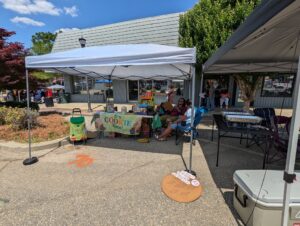 Outdoor market stand with a banner reading "It's Cookie Time!", people sitting under tents, and signs for cookies and peanut butter sandwiches.