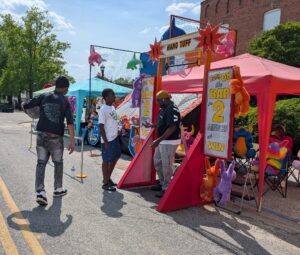 People at a carnival game booth with colorful stuffed animal prizes, signs reading "Hang on the bar 2 minutes and win," and a bright sunny day.