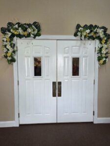 White double doors with rectangular windows, decorated above with green foliage and yellow-white floral arrangements, against a beige wall and brown carpeted floor.
