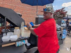 Man in a red shirt grilling food outside, standing by a large barbecue smoker, under an umbrella, with supplies and containers on a side shelf.
