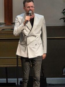 A man wearing glasses and a light-colored blazer speaks into a microphone while standing indoors.