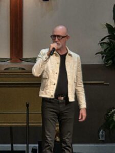 A bald man with glasses sings into a microphone, wearing a white jacket and black pants, standing indoors near a wooden cross and some plants.