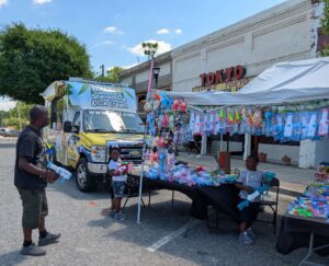 Man and two children at an outdoor toy stand near a shaved ice truck, with stuffed animals and toys displayed under a canopy.