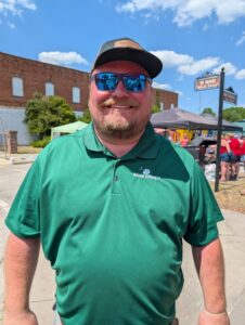 Man in a green Dillon County shirt, sunglasses, and cap, smiling outdoors at a sunny street event with tents and people in the background.