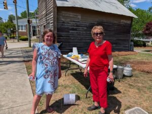Two women standing outside near a rustic wooden building, smiling at the camera, with a table of items and supplies behind them on a sunny day.