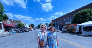 Two women stand smiling on a closed street during a sunny outdoor market event, with food trucks and vendors lining both sides of the road.