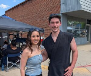 A woman and man smile outdoors; she wears a strapless top, he wears a black martial arts uniform. Tents and people are visible in the background.