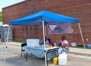 Two people sit under a blue canopy at a street vendor booth, selling baked goods from "Millz Bakery" with a sign and coolers on the table.