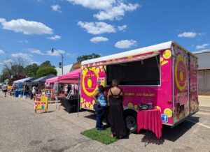 People standing at a bright pink lemonade food truck at an outdoor market with tents and vendors on a sunny day.