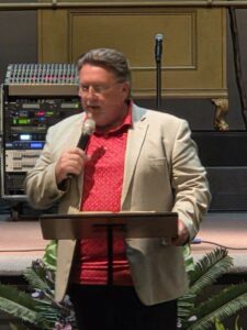 A man in a beige blazer and red shirt speaks into a microphone at a podium, with audio equipment visible in the background.