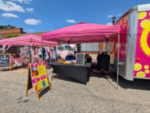 Outdoor market with pink tents, a food truck, clothing racks, a chalkboard menu, and a sign listing drink specials on a sunny day.