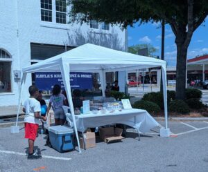People gather at a white tented booth on a sunny day, selling baked goods. A blue banner reads "Kayla's Kreationz & More." Children stand nearby holding a balloon.