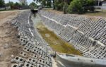 Concrete drainage ditch lined with perforated blocks, containing a small amount of water, surrounded by grass and trees under a clear sky.