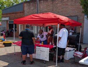 People at a street food stall under a red canopy, selling barbecue and various foods, with a handwritten menu and grill visible, in an outdoor urban setting.