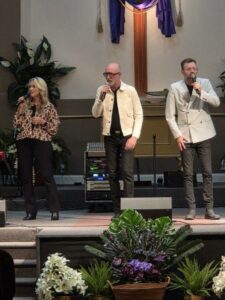 Three people sing on a church stage with microphones, standing in front of a cross, surrounded by plants and floral arrangements.