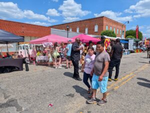 People walk and gather at a lively outdoor street market with colorful tents, food trucks, and a pink VW van under a sunny, blue sky with scattered clouds.