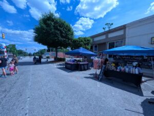 Street market with vendor tents, clothing displays, beauty products, and people walking on a sunny day with blue sky and scattered clouds.