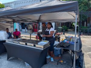 People serving food under a canopy at an outdoor market, with trays of food on tables and two women seated in the background.