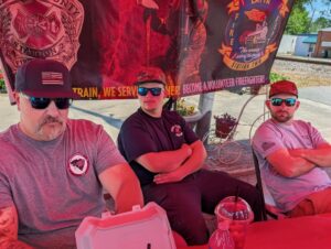 Three men wearing sunglasses and firefighter shirts sit at a table under a tent, promoting volunteer firefighter recruitment. Banners and drinks are visible on and around the table.