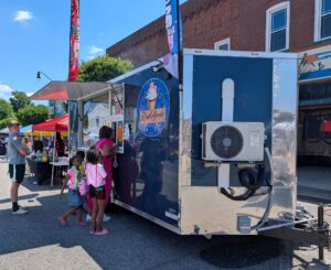 People stand in line at a blue "Royal Sweets" food truck at an outdoor event, with tents and buildings visible in the background.