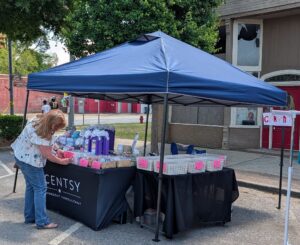 A woman arranges products under a blue canopy at an outdoor clearance sale, featuring pink sale signs, on a city sidewalk near a building with red doors.