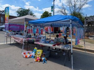 Outdoor market stalls with toys, plushies, and treats, including an Italian Ice vendor, colorful banners, and two people sitting under a blue canopy on a sunny day.