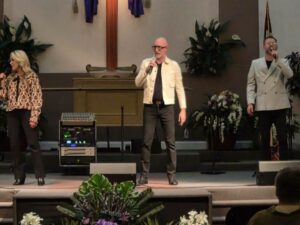 Three people singing on stage in a church, surrounded by plants and flowers, with a wooden cross and sound equipment behind them.