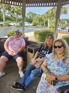 Three people sitting on benches under a gazebo, smiling at the camera, with palm trees and parked cars visible in the background on a sunny day.