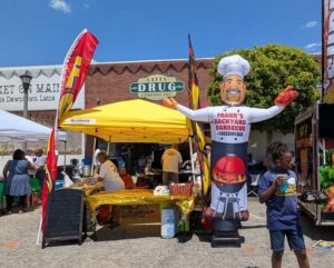 Outdoor barbecue stand with a yellow tent, inflatable chef advertising "Frank's Backyard Barbecue," people browsing food, and a child with a drink at a street fair.