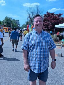 A man in a blue plaid shirt stands smiling at an outdoor street fair, with people, booths, and inflatables in the background on a sunny day.