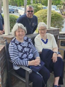 Two elderly women sit and smile outdoors on a bench, while a man in sunglasses stands behind them. It appears to be a sunny day with greenery and cars in the background.