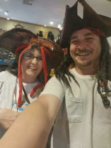 Two people smiling and wearing pirate hats, one with fake dreadlocks and beads, posing for a selfie indoors.