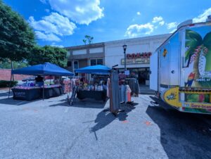 Outdoor market with blue tents, tables displaying products, clothing racks, and a colorful truck. A restaurant called "Tokyo" is in the background under a partly cloudy sky.