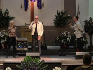Three people singing with microphones on a church stage, surrounded by flowers and plants, with a cross and sound equipment in the background.