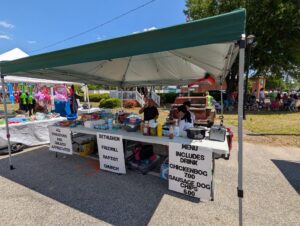 Outdoor church food stall under a canopy, serving chicken bog and sausage dogs with drinks and chips, donation signs displayed, toys and other items for sale nearby.