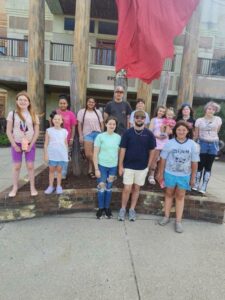 A group of people, including children and adults, stand together outside in front of large wooden poles and a red flag, posing for a photo.