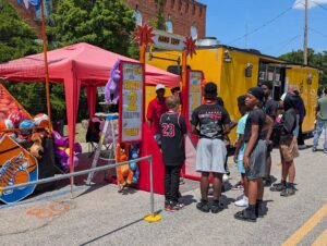 A group of kids stand in line at a carnival booth with stuffed animal prizes and a challenge to hang on a bar for two minutes.