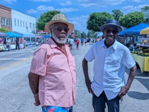 Two men wearing hats and sunglasses stand on a street at an outdoor market or festival, with vendor tents and people in the background.