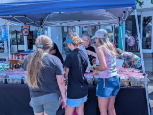 Three people shopping at an outdoor market stall with baked goods under a blue canopy, interacting with the vendor on a sunny day.