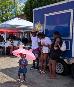 A young girl holds a pink umbrella while three adults stand near a food truck at an outdoor event on a sunny day.